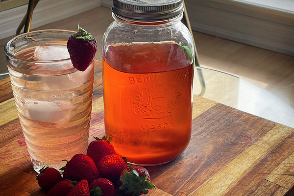 A tall glass of pale pink cocktail with a strawberry on the rim beside a mason jar of deep pink-orange shrub