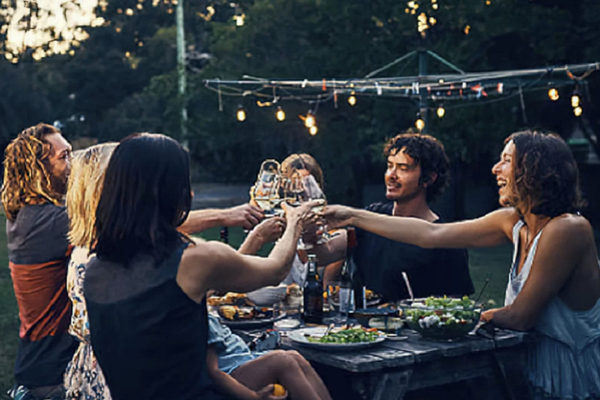 A group of men and women sitting around an outdoor table in a garden under a string of lights, laughing and clinking wine glasses