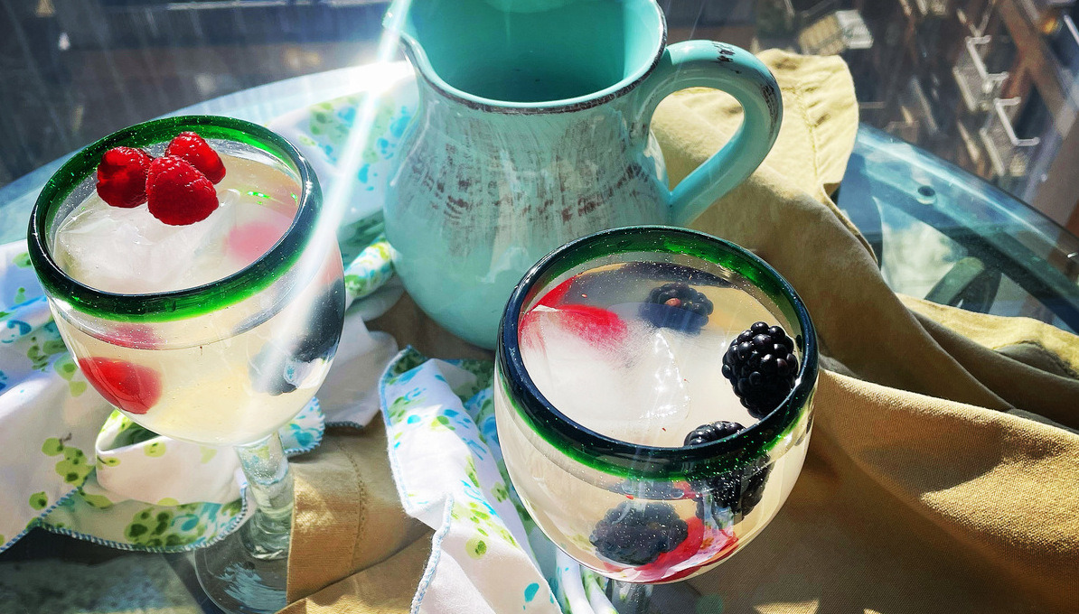 An outdoor table on a sunny day with a turquoise ceramic pitcher flanked by two Mexican-styled wine glasses filled with pale sangria and berries floating in each
