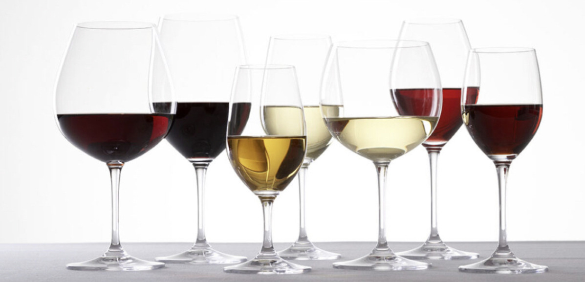 7 wine glasses of various heights and shapes, filled with different coloured wines, lined up against a glowing white wall