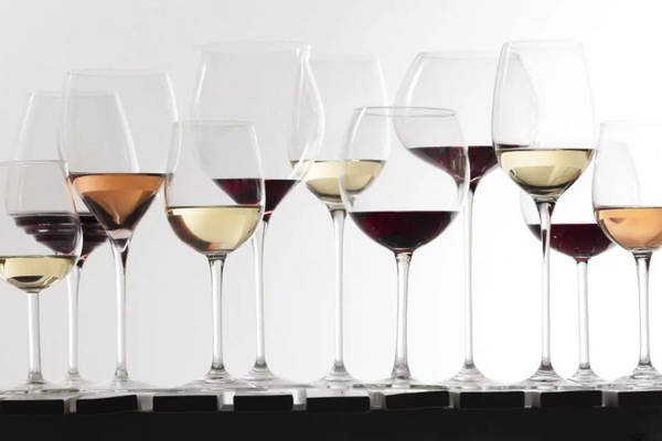 11 wine glasses of various heights and shapes, filled with different coloured wines, lined up against a glowing white wall