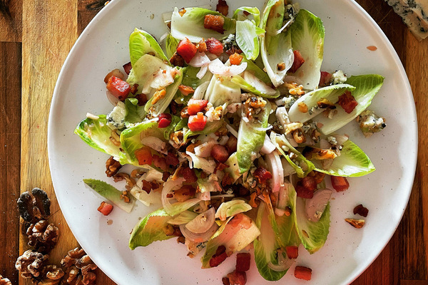 overhead view of a mixed salad on a white plate placed on a wooden board. Beside the plate is crumbled blue cheese in the top right corner and walnut pieces in the bottom left.