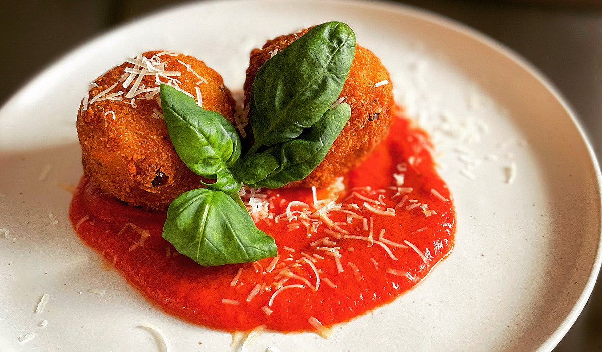 A white plate with two, round arancini in a puddle of tomato sauce with a basil leaf