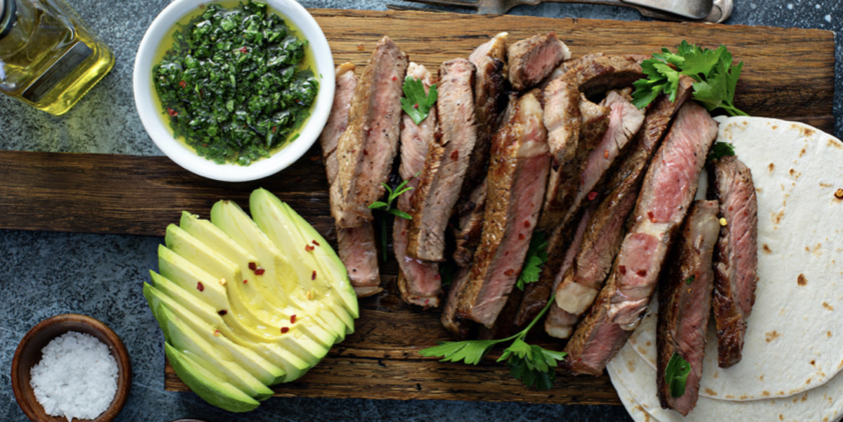 A wooden platter with tortilla shells and slices of steak with a bowl of green chimichuri sauce