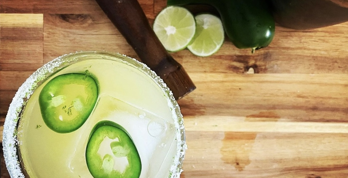 Overhead shot of a margarita in a salt rimmed glass with 2 slices of jalepeno floating in it, sitting on a wooden board. Beside the drink is a muddler, jalepeno and cut lime.