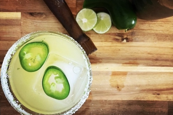 Overhead shot of a margarita in a salt rimmed glass with 2 slices of jalepeno floating in it, sitting on a wooden board. Beside the drink is a muddler, jalepeno and cut lime.