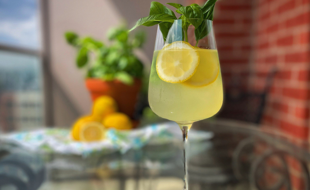 A limoncello spritz in a wine glass with lemon wheels and basil leaves sits on a glass table outdoors. A basil plants and lemons are blurry in the backgroun