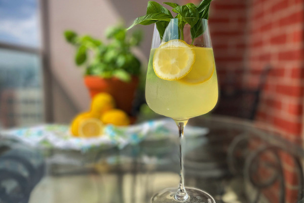 A limoncello spritz in a wine glass with lemon wheels and basil leaves sits on a glass table outdoors. A basil plants and lemons are blurry in the background.