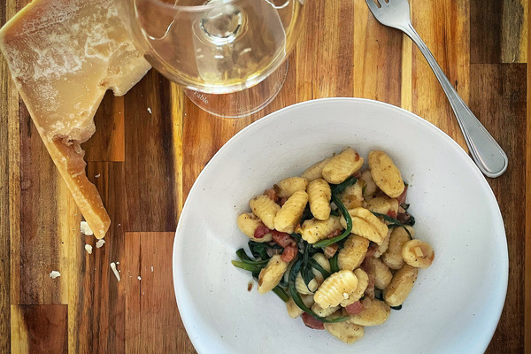 A bowl of gnocchi on a wood board, green ramps laced throughout, a glass of white wine beside the bowl and a wedge of parmesan beside it.