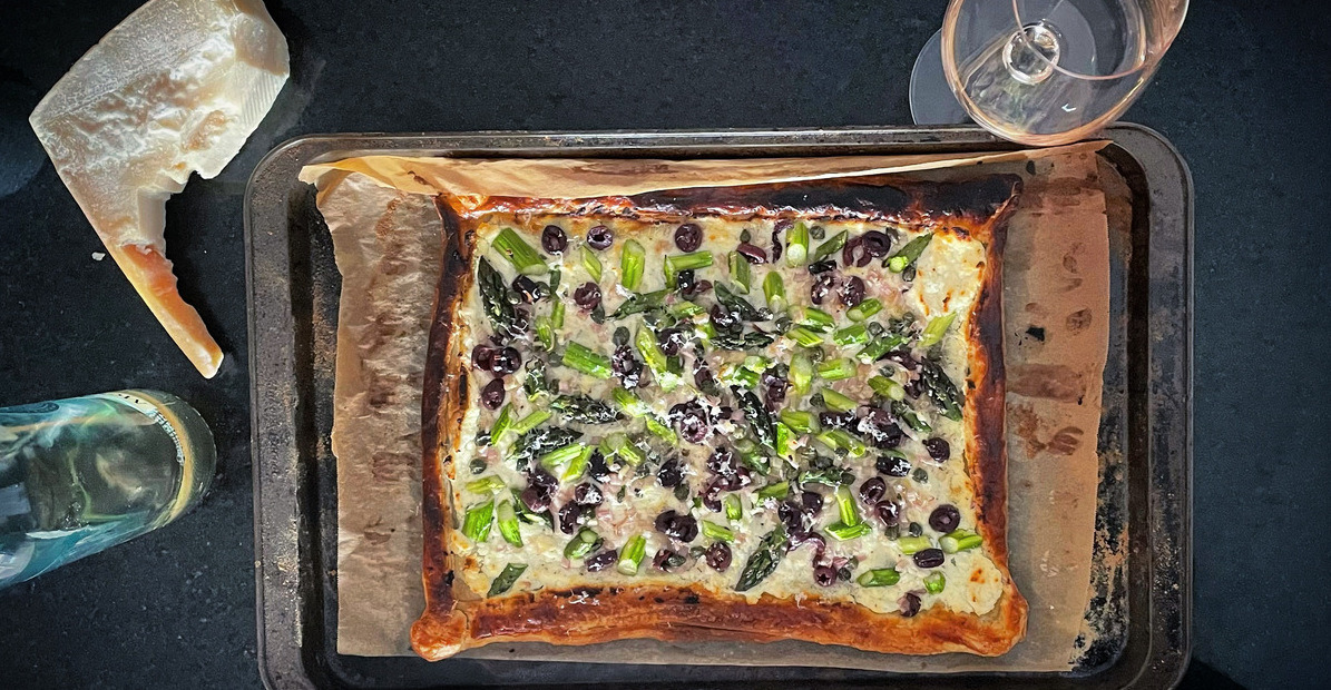 Overhead view of rectangular tart filled with asparagus, purple olives and cheese, on a baking tray sitting on a black counter with a wedge of cheese and glass of rosé on either side.