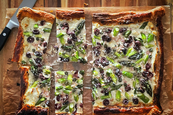 Overhead view of rectangular tart filled with asparagus, purple olives and cheese, on a wood board with a sharp knife to the side