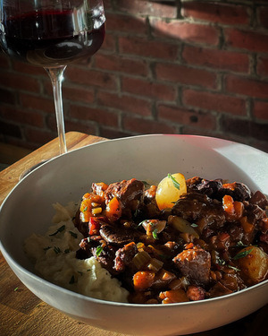 a white bowl of boeuf bourguignon in the setting sun with a glass of red wine in front of a brick wall