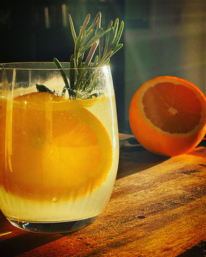 A glass tumbler with clear liquid, an orange wheel and sprig of rosemary sit on a wooden cutting board, illuminated by sunlight. Behind the glass is a half cut orange and pairing knife with water drops on it.