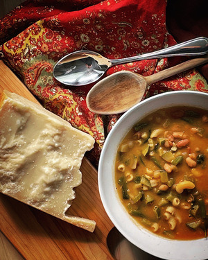 a white bowl filled with hearty minestrone. beside the bowl is a hunk of crumbly parmesan on a wood board and a rust coloured napkin with a wood spoon