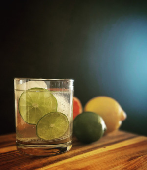 Clear cocktail in a rock glass filled with ice and two rounds of lime floating. The glass is set against a black background and mixed citrus fruit lie behind it.