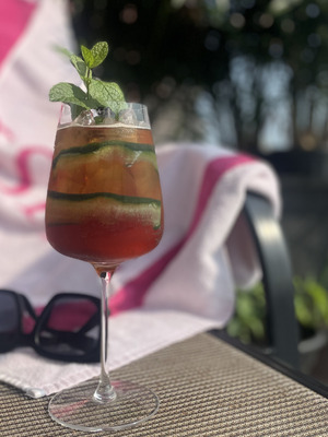 A Pimm's cup, with mint and cucumber, sitting on a lawn chair with a beach towel and sunglasses in the background
