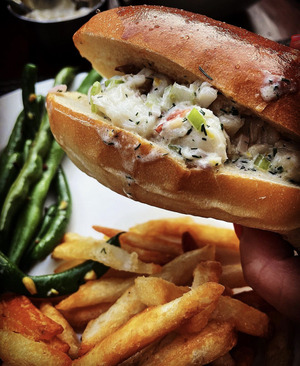 A close up of a woman's hand holding a lobster roll, in the background is a plate of salad and fries.