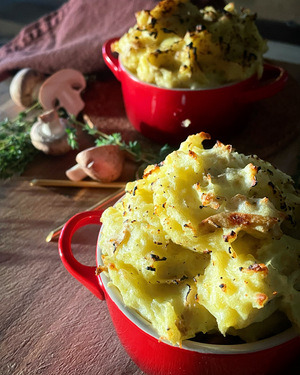 Two red ramekins topped with crusty mashed potato on a wooden board with scattered thyme and mushrooms